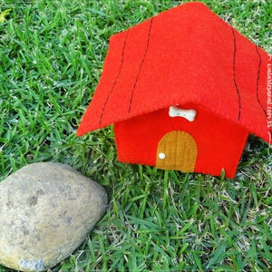 Op de afbeelding: Een rood vilten hondenhuis met een bruine vilten deur en een wit bot op het dak. Het hondenhuis staat op groen gras.