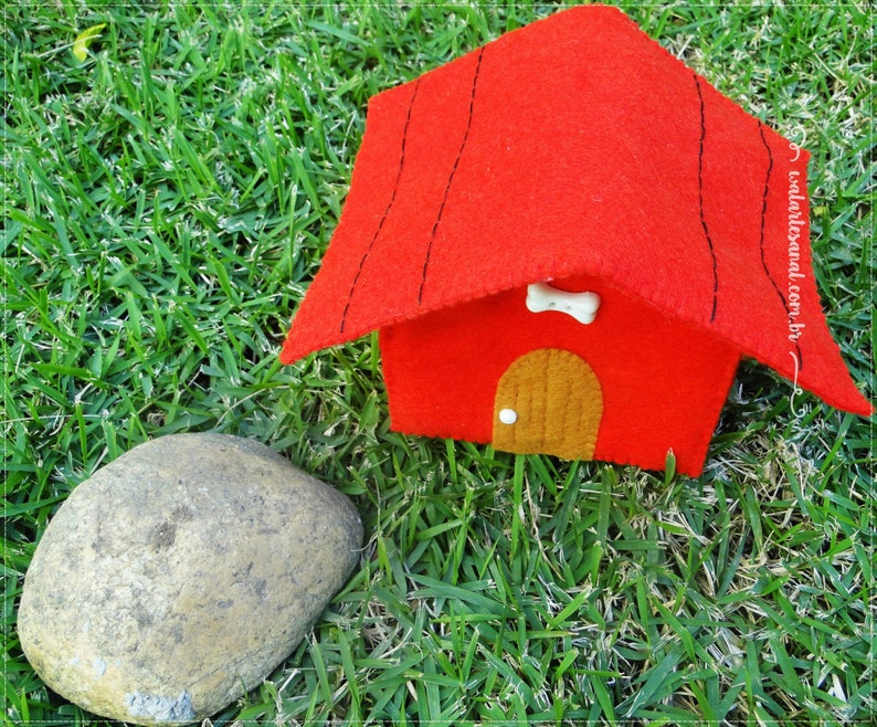 Op de afbeelding: Een rood vilten hondenhuis met een bruine vilten deur en een wit bot op het dak. Het hondenhuis staat op groen gras.