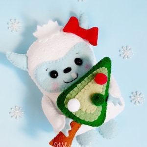 May include: A white felt snowman with a blue face, holding a green felt Christmas tree with red and white felt ornaments. The snowman is wearing a red bow.
