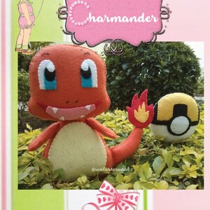 Könnte beinhalten: Ein Charmander Plüschtier aus Filz mit einem roten und orangen Körper, einer gelben Schwanzflamme und einem weißen Pokéball. Das Spielzeug sitzt auf einem Bett aus grünen Blättern.