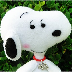Op de afbeelding: Witte pluche speelgoed van Snoopy, een cartoon beagle, met zwarte vilt ogen, neus en oren. Snoopy draagt een rode vilt kraag met een zilveren hanger.