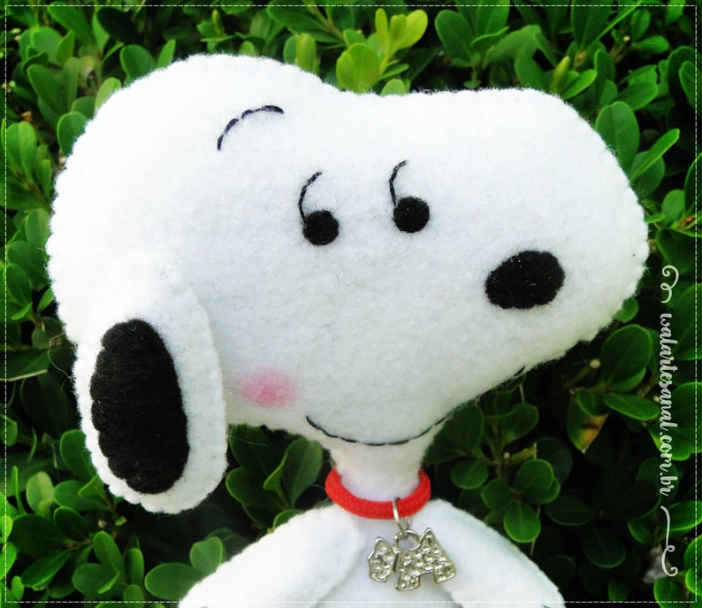 Op de afbeelding: Witte pluche speelgoed van Snoopy, een cartoon beagle, met zwarte vilt ogen, neus en oren. Snoopy draagt een rode vilt kraag met een zilveren hanger.