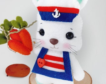 May include: A white felt bunny wearing a blue and red sailor hat with an anchor, a red and white striped shirt, and holding a carrot. The bunny is standing on a wooden slice.
