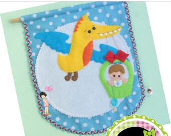 May include: A felt wall hanging with a blue background and white polka dots. A yellow dinosaur with blue wings is carrying a baby in a green basket. The text "Boy and the Dinosaur" and "O Menino e o Dino" is at the top of the hanging.