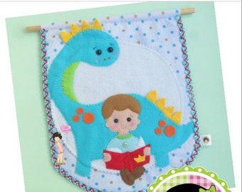 May include: A blue felt dinosaur with a yellow crest and orange spots, holding a red book. A boy with brown hair and a blue shirt is sitting on the dinosaur's back. The background is a white felt with brown and blue polka dots. The text "*BOY AND THE DINOSAUR*" and "★Ο ΜΕΝΙΝΟ E O DINO ★" is at the top of the image. The text "Wal Artesanal" and "Copyright 2020 - Wal Vieira" is at the bottom of the image.