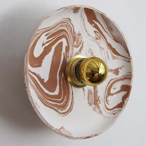 Peut inclure: Applique murale ronde, blanche et marron marbrée, avec un luminaire de couleur laiton. L'applique a une forme peu profonde en forme de plat avec un motif tourbillonnant de marron et de blanc. Le luminaire a une ampoule ronde de couleur or.