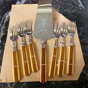May include: A vintage silverware set featuring a cake server and six forks. The cake server and forks have stainless steel heads and handles with alternating light and dark wood grain. The set is displayed on a cardboard backing.