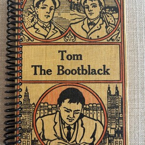 May include: A spiral-bound notebook with a vintage illustration on the cover. The cover features portraits of young men and the title "Tom The Bootblack." The illustrations are in shades of orange, brown, and black, with a textured background.