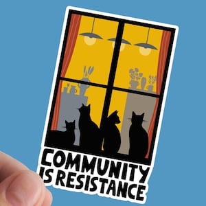 May include: A sticker with a window scene of four black cats looking out of a window with yellow curtains. The text below the window reads "Community is Resistance."