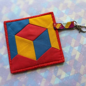 May include: A handmade, square quilted coaster with a geometric design in red, yellow, and blue. The coaster has a red border and a small fabric loop with a metal clasp attached. The design resembles a 3D cube.