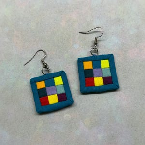May include: A pair of square earrings featuring a teal fabric border surrounding a grid of nine small, multicolored fabric squares in shades of yellow, lime green, light blue, dark blue, lavender, red, and deep purple, attached to silver-tone hooks.