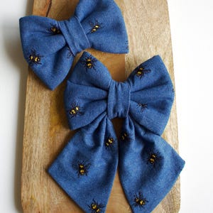 Pode incluir: Dois laços de cabelo de tecido jeans azul com um padrão de abelhas. O laço maior é amarrado com uma cauda longa.