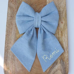 May include: A large, light blue fabric bow with long tails, resting on a wooden surface. The bow is tied in the center and has the name "Remi" embroidered in a delicate, cursive font on one of the tails.