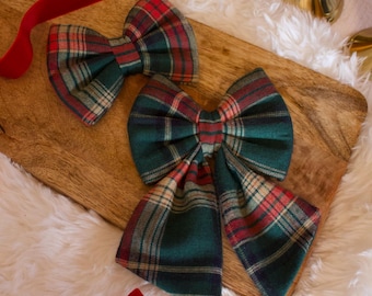 May include: Two plaid fabric bows in green, red, and cream, with a red velvet ribbon, rest on a wooden cutting board. The bows are on a fluffy white surface, with gold candle holders in the background. Festive holiday decor.