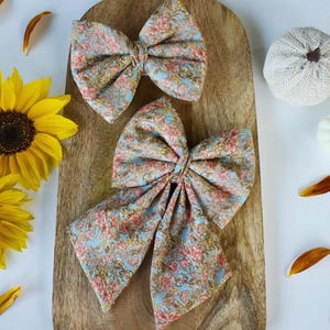 Può includere: Due fiocchi per capelli in tessuto floreale azzurro, rosa e giallo, su un tagliere di legno. Girasoli, petali e zucche decorative circondano i fiocchi. I fiocchi misurano circa 15 cm.