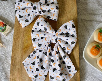 May include: Two white fabric hair bows with a repeating pattern of black cat faces, orange crescent moons, and stars. The bows are on a wooden chopping board. The bows are likely for Halloween.