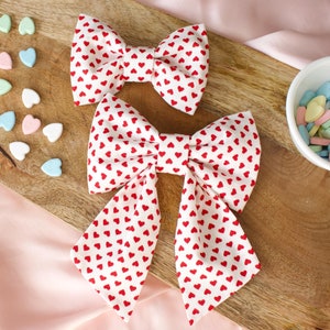 May include: Two white fabric bows with red heart patterns. The larger bow is tied in a loop with the ends hanging down. The smaller bow is tied in a knot.