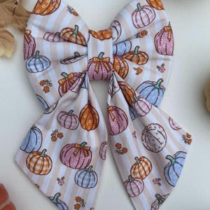May include: A fabric hair bow featuring a repeating pattern of pumpkins in shades of orange, pink, and blue, interspersed with small orange flowers on a background of vertical beige and white stripes. The bow has two long, pointed tails.