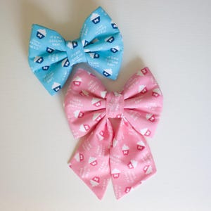 May include: Two fabric bow ties, one blue and one pink, with a white cupcake and text pattern. The text on the bows reads "It's my Gotcha Day!"
