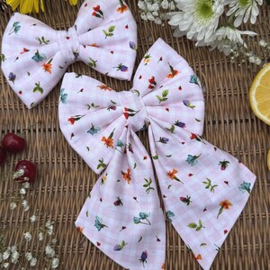 Puede incluir: Dos lazos para el pelo de cuadros rosas con un estampado floral. Los lazos presentan un patrón repetido de pequeñas flores en tonos naranja, azul y morado, con hojas verdes. Los lazos están colocados sobre una superficie tejida.
