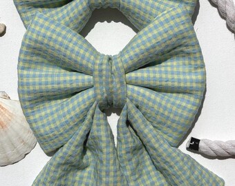 May include: Two hair bows made of light blue and pale yellow gingham fabric. The top bow is a standard shape, while the bottom bow features two long, pointed tails extending downward. Both bows have a gathered center knot made of the same fabric.