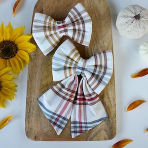 May include: Two cream, red, brown, and blue plaid fabric hair bows. The bows are displayed on a wooden board with sunflowers and decorative pumpkins. The bows are in a classic bow shape.