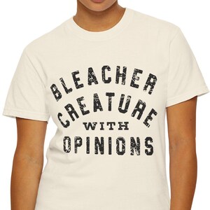 May include: Cream-colored t-shirt with the text "BLEACHER CREATURE WITH OPINIONS" in a distressed black font. The shirt is a crew neck and is being worn by a person wearing blue jeans.