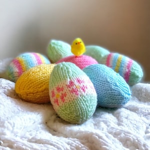 May include: A group of knitted Easter eggs in various colors, including yellow, pink, blue, and green, with a small yellow chick on top of one of the eggs. The eggs are sitting on a white fluffy surface.