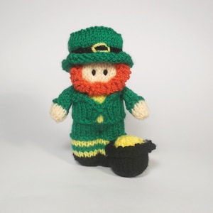 May include: A knitted leprechaun doll. It features a green hat, jacket, and trousers, with an orange beard. The doll has a yellow shirt and holds a black pot filled with gold coins. A festive decoration.