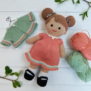 May include: A knitted doll with brown hair in two buns, wearing a peach dress with a white collar and black shoes with white socks. Beside the doll is a matching green knitted cardigan on a wire hanger and two balls of yarn in peach and green.
