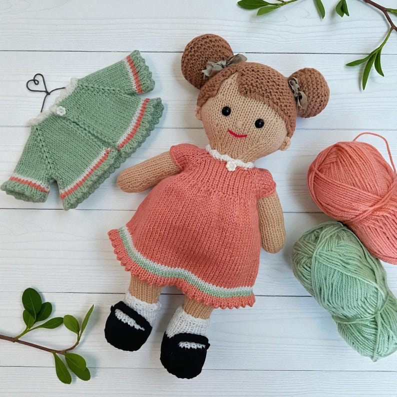 May include: A knitted doll with brown hair in two buns, wearing a peach dress with a white collar and black shoes with white socks. Beside the doll is a matching green knitted cardigan on a wire hanger and two balls of yarn in peach and green.