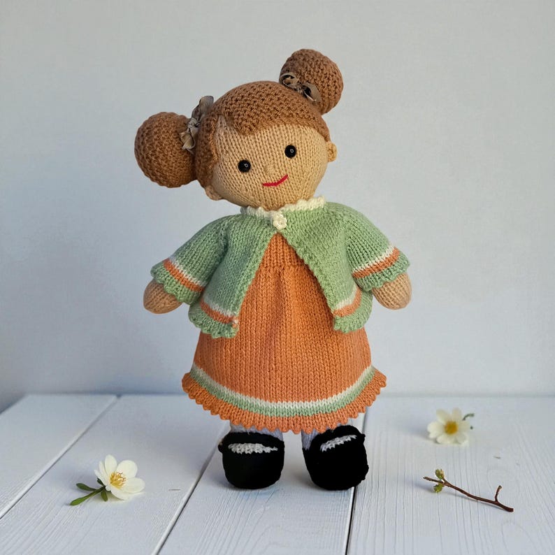 May include: A handcrafted knitted doll with brown yarn hair styled in two buns with small bows. The doll wears a light green cardigan with orange and white striped cuffs over an orange dress with a white hem. It has black shoes and white socks.