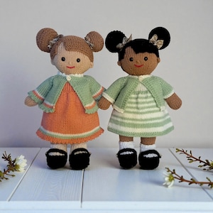Op de afbeelding: Twee gebreide poppen met garen haar in knotjes met kleine strikjes. De linkerpop heeft lichtbruin haar, een oranje jurk en een saliegroen vestje. De rechterpop heeft zwart haar, een wit en saliegroen gestreepte jurk en een saliegroen vestje.