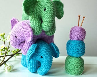 Op de afbeelding: Drie handgebreide olifantjes in groen, paars en blauw, verticaal op elkaar gestapeld. Daarnaast staat een stapel van drie bollen wol in bijpassende kleuren, bekroond met twee houten breinaalden. De olifantjes hebben kleine zwarte kraaloogjes.