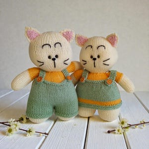 May include: Two handmade knitted cat dolls with cream-colored yarn bodies and pink inner ears. One cat wears sage green overalls with orange buttons, while the other wears a matching sage green pinafore dress with an orange hem and buttons.