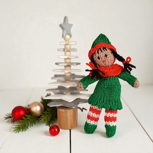 May include: A hand-knitted doll featuring a green dress, a red scarf, and a matching green hat. The doll has dark brown yarn hair styled in two pigtails and wears red and white striped leggings. It stands beside a wooden star-shaped tree.