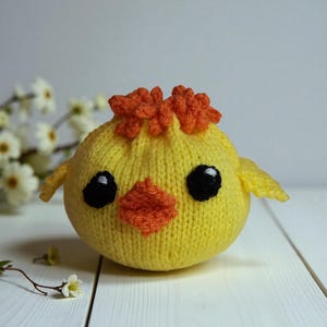 May include: A round, yellow knitted chick toy featuring a textured orange tuft on top, a small orange beak, and two large black eyes with white highlights. The soft, spherical body has small knitted wings on each side, creating a simple, plush form.