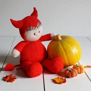 Puede incluir: Una muñeca tejida con un disfraz de diablo rojo con cuernos y cola, con cara color crema y cabello castaño. La muñeca está sentada junto a una calabaza amarilla grande y dos calabazas tejidas pequeñas de color naranja y hojas otoñales.