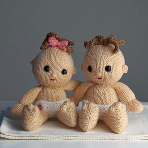 May include: Two handmade knitted dolls with light beige yarn skin, black bead eyes, and small red stitched mouths. Both dolls wear white knitted diapers. One doll features brown curly hair with a small pink bow, while the other has brown curly hair.