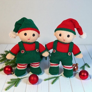 May include: Two handmade knitted dolls with beige skin, black eyes, and red mouths. One doll wears a green hat with a white pom-pom and a green dress over a red shirt. The other wears a red hat with a white pom-pom and green overalls over a red shirt.