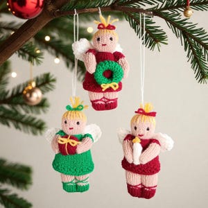 May include: Three knitted angel ornaments hanging from white strings. Each angel has blonde hair, white wings, and pink skin. One wears a red dress holding a green wreath, one a green dress holding a red gift, and one a red dress holding a white candle.