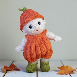 May include: A hand-knitted doll featuring a cream-coloured body, wearing a ribbed orange pumpkin-shaped hat with a small green stem on top, a matching orange pumpkin-style romper, and solid green booties. The doll has black bead eyes and a pink mouth.