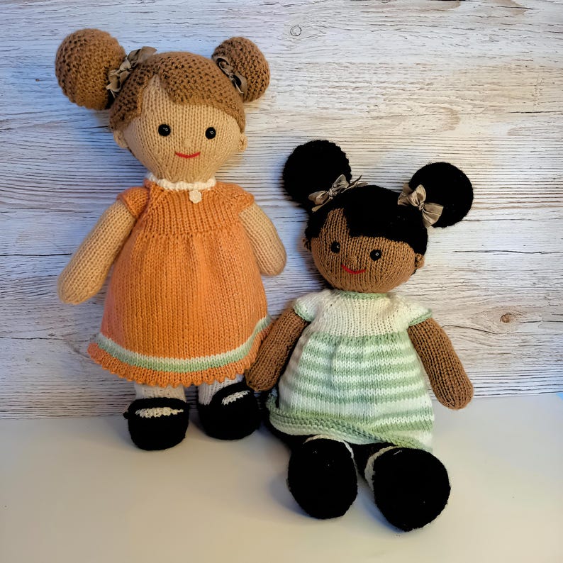 May include: Two handmade knitted dolls with yarn hair styled in double buns with small ribbons. The taller doll has light brown skin and wears an orange dress with a light green hem. The shorter doll has dark brown skin and wears a green and white striped dress. Both dolls have black shoes.