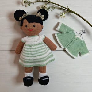 May include: A knitted doll with dark skin and black hair styled in two buns with small beige bows. The doll wears a white and light green striped dress, white socks, and black shoes. A separate light green knitted cardigan is included.