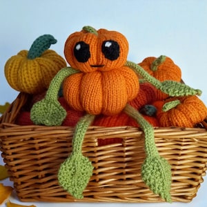 May include: A knitted orange pumpkin character with large black eyes and a small mouth, featuring green vine-like arms and legs with leaf-shaped hands and feet, sitting inside a woven wicker basket filled with several other knitted pumpkins.