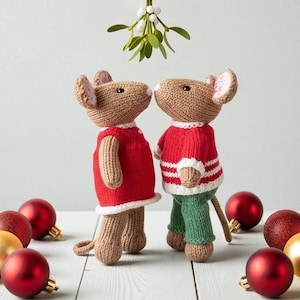 May include: Two knitted mice, nose to nose, under mistletoe. One mouse wears a red dress with white trim, the other a red sweater with white stripes and green pants. Red and gold Christmas ornaments are scattered around.