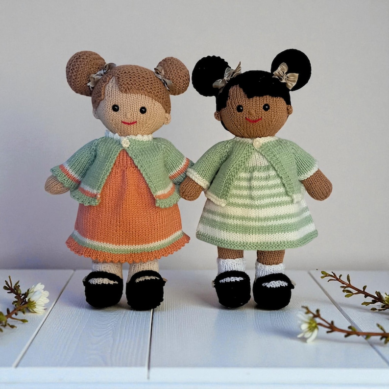 May include: Two knitted dolls with yarn hair styled in buns with small bows. The left doll has light brown hair, an orange dress, and a sage green cardigan. The right doll has black hair, a white and sage green striped dress, and a sage green cardigan.