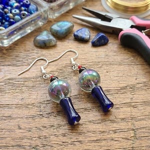 May include: A pair of handmade earrings with iridescent glass orbs and dark blue glass stems. The earrings are suspended from silver-colored hooks. Beads and jewelry-making supplies are in the background.