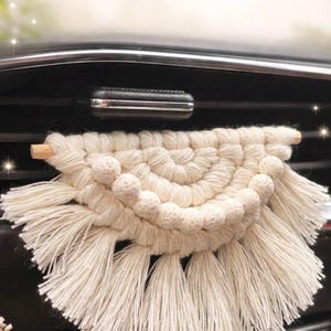 Könnte beinhalten: Cremefarbene Makramee-Dekoration für die Lüftung im Auto. Das handgefertigte Stück hat ein halbkreisförmiges Design mit geknoteten Details, kleinen Kugelakzenten und Fransen. Es ist an einem Holzstab befestigt.