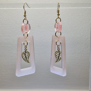 Op de afbeelding: Een paar hangende oorbellen met een roze en gouden ontwerp. De oorbellen hebben een rechthoekig frame met een gouden blaadjesbedel die erin hangt. Het frame is bevestigd aan een roze kraal en een gouden haak.